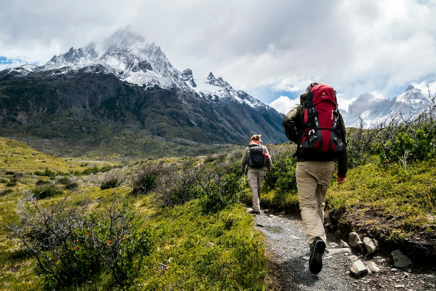 Two hikers on a trail walking toward snowy mountain peaks